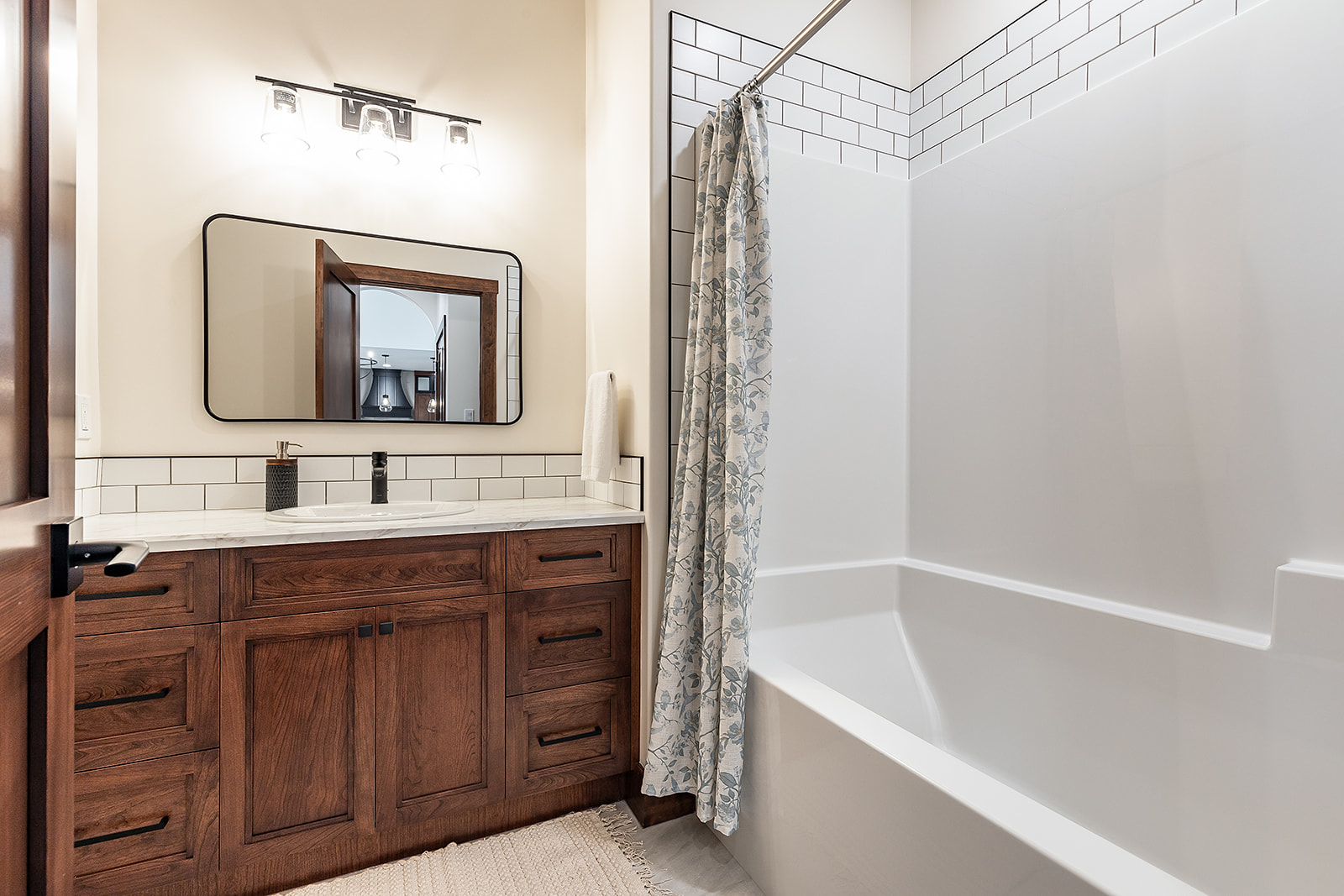 Modern bathroom featuring a wooden vanity with white countertop, large mirror, and three-light fixture. A bathtub with a patterned shower curtain is on the right.