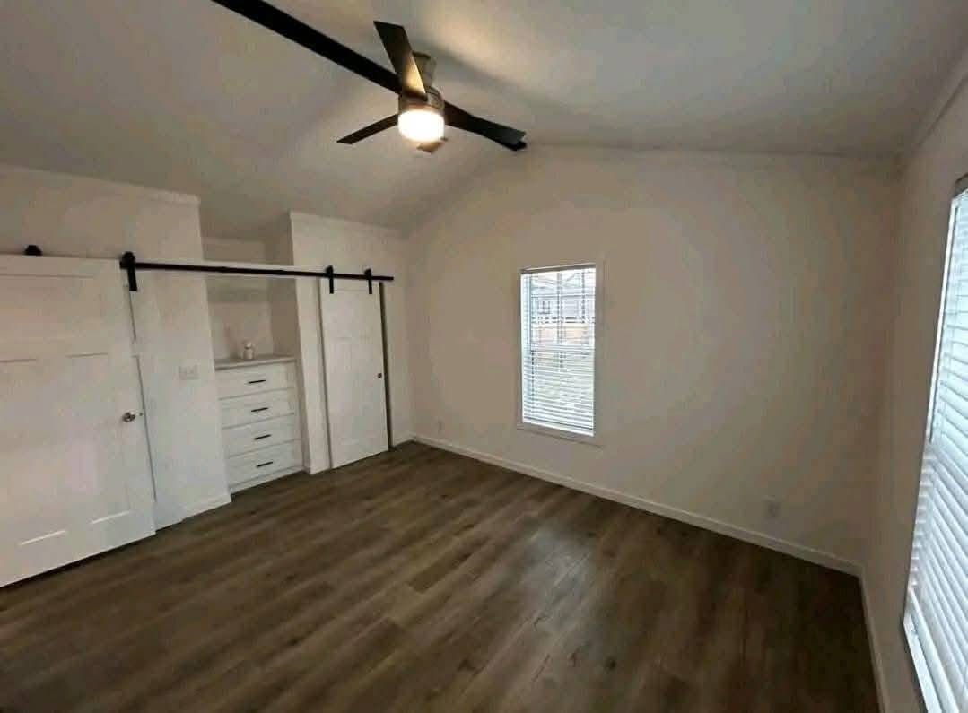 A spacious, empty room with white walls and brown wood flooring. Features a ceiling fan, a window with blinds, and a sliding barn door revealing shelves.