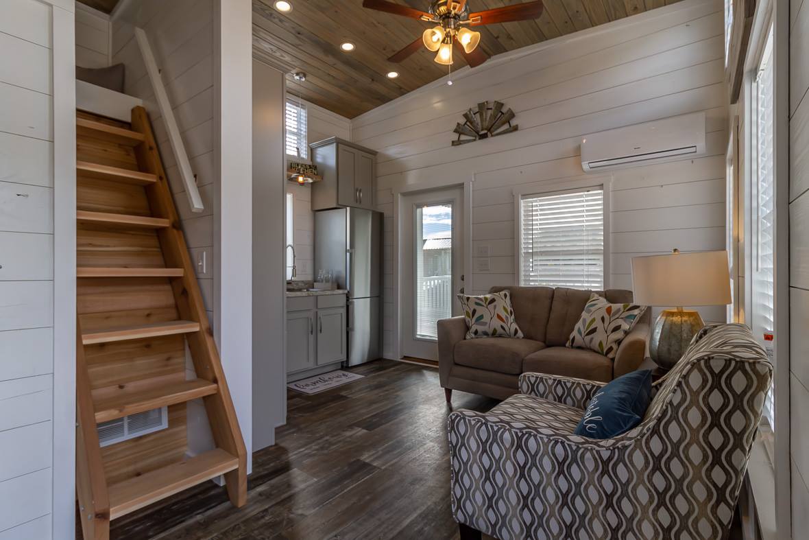 Cozy tiny home interior with wooden stairs, compact kitchen, stylish living area featuring a patterned chair, sofa, lamp, and ceiling fan. Bright and warm.