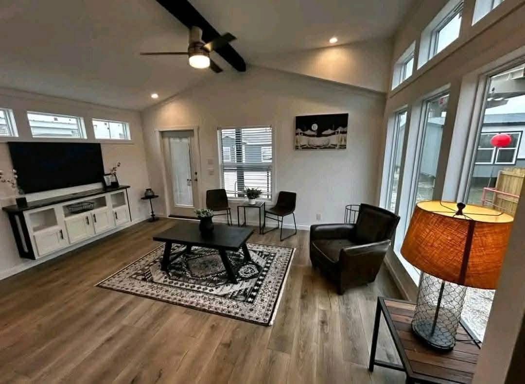 Modern living room with wooden floors, large windows, a sleek ceiling fan, cozy seating, and a flat-screen TV. Warm lighting creates a welcoming atmosphere.