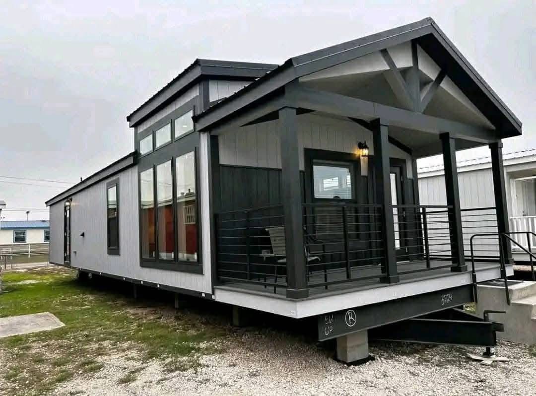Modern mobile home with large windows and covered porch on a gravel base. The exterior features gray siding and dark trim, evoking a cozy, inviting atmosphere.