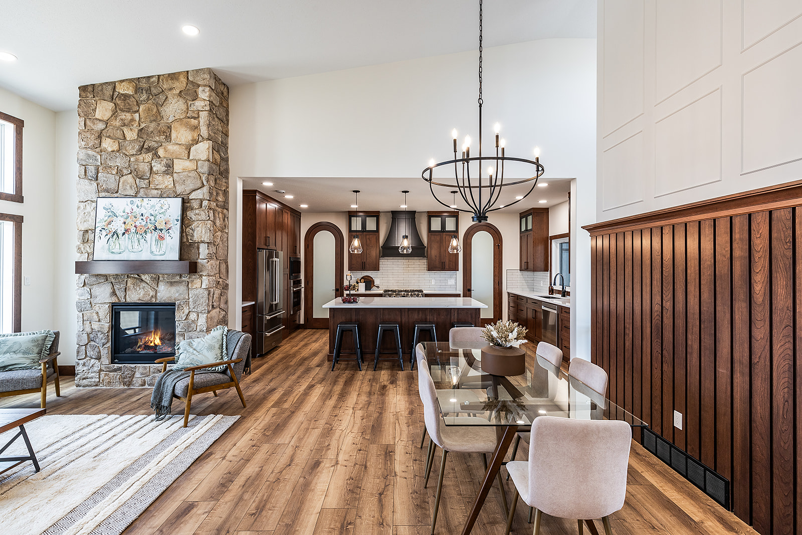Sophisticated open-concept living space, featuring a stone fireplace, elegant chandelier, wood floors, and a modern kitchen with dark cabinets. Cozy, inviting ambiance.