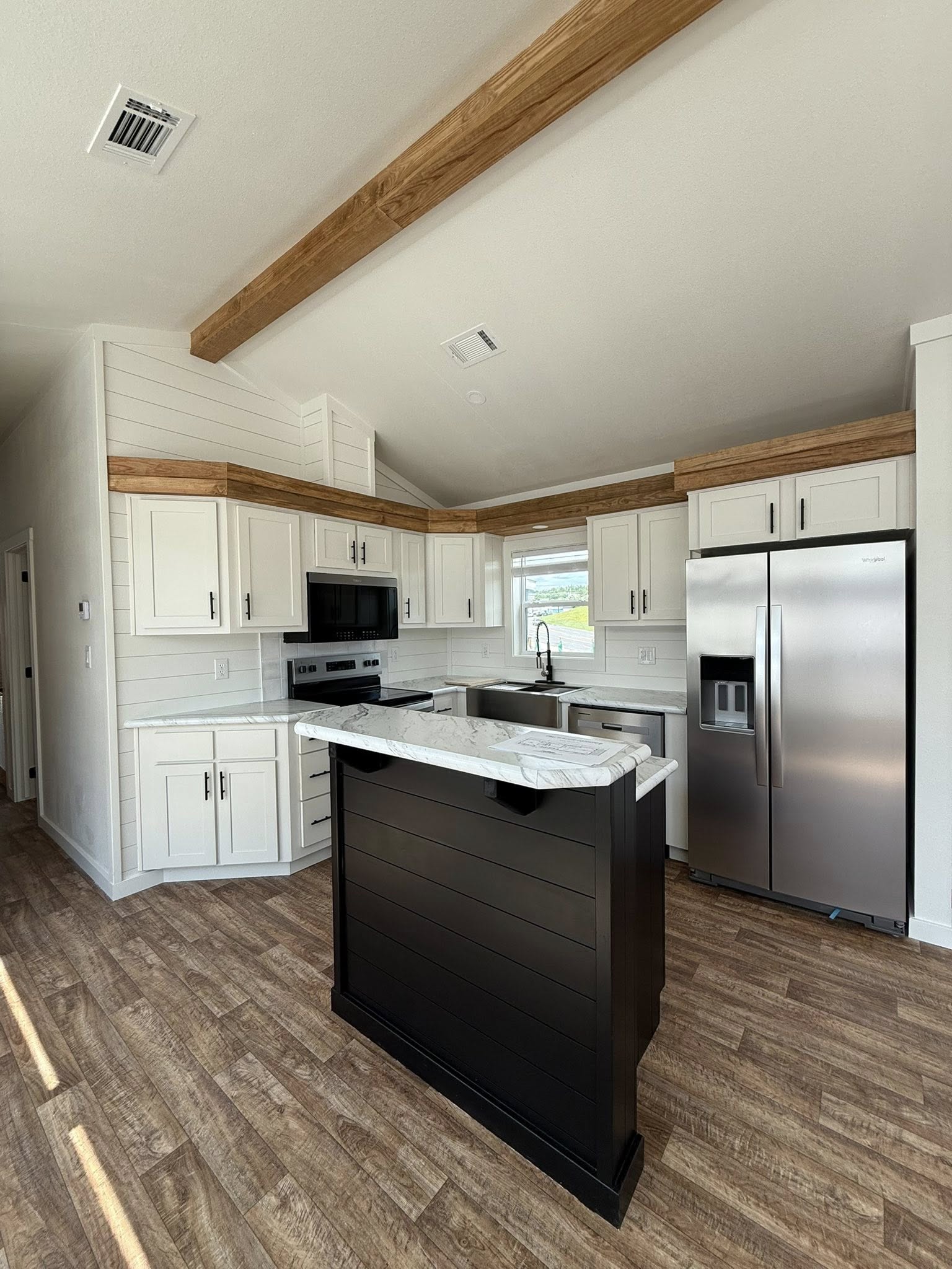 Modern kitchen with light wood floors, a vaulted ceiling with wooden beams, white cabinets, stainless steel appliances, and a black island with marble countertop.