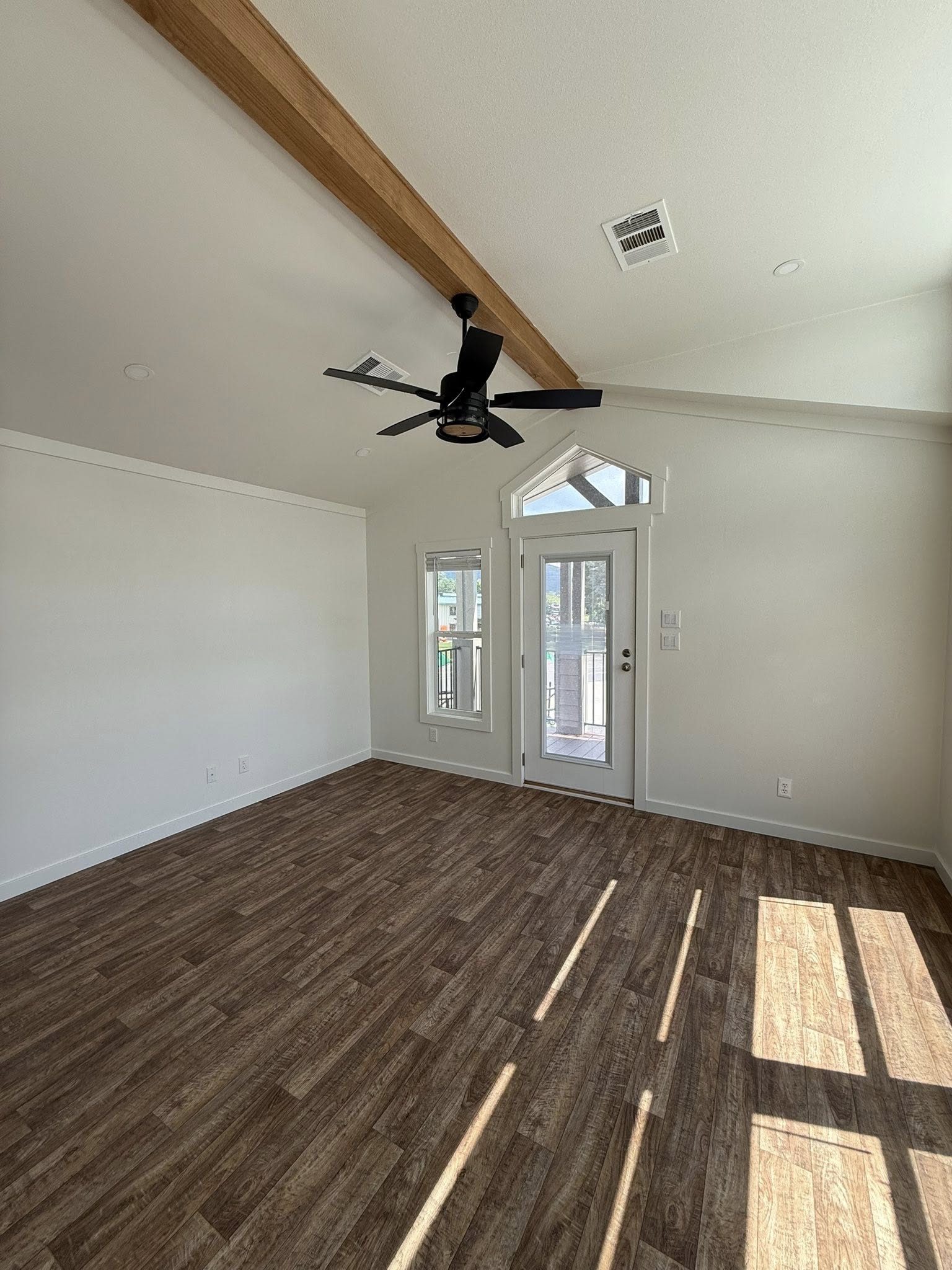 Spacious room with vaulted ceiling, wooden beam, and ceiling fan. Sunlit wood floor, white walls, and a glass door with sidelights create a bright atmosphere.