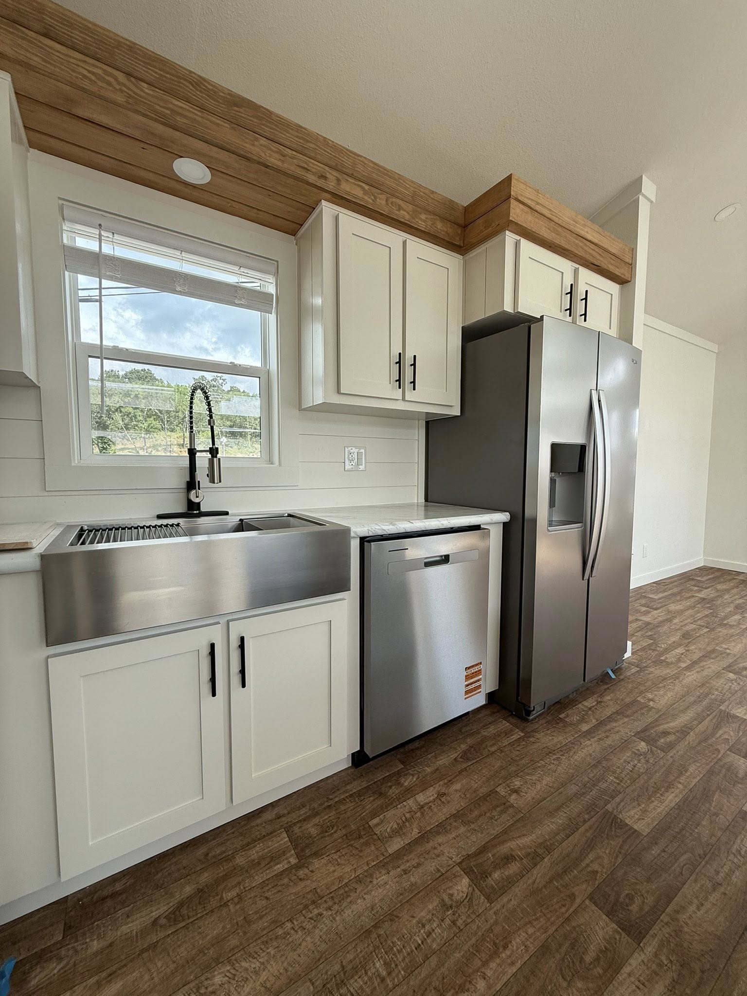 A modern kitchen with white cabinets, stainless steel appliances including a large fridge, farmhouse sink, and dishwasher. Wooden floor and serene view.