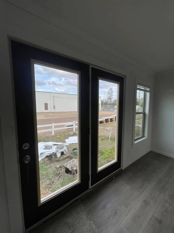 View from inside a room with dark-framed glass doors and a window, showing a grassy area outside, white fence, and an industrial building under a cloudy sky.