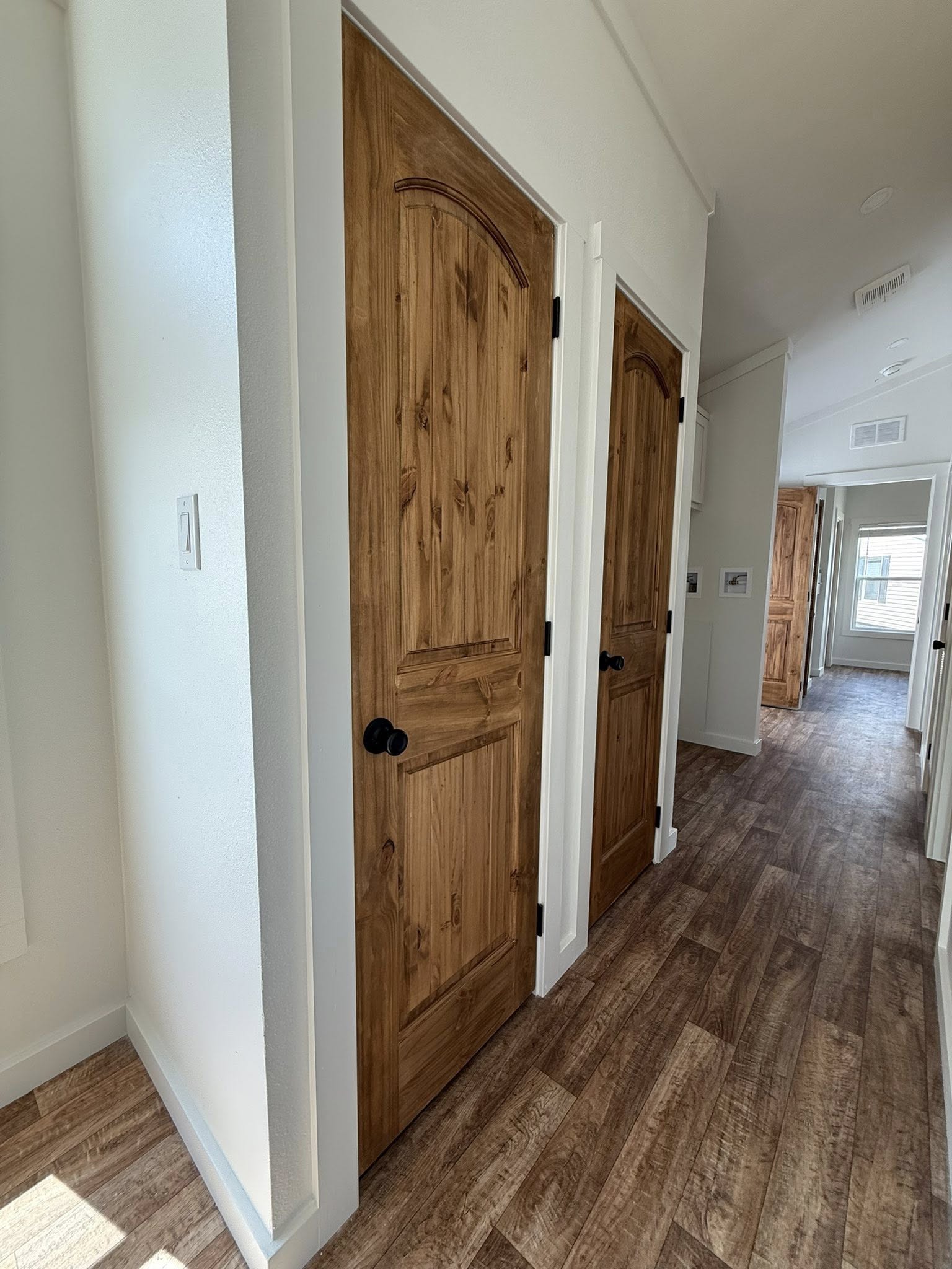 A hallway with wooden flooring features two closed wooden doors on the left, leading to an airy room with sunlight streaming through a window.