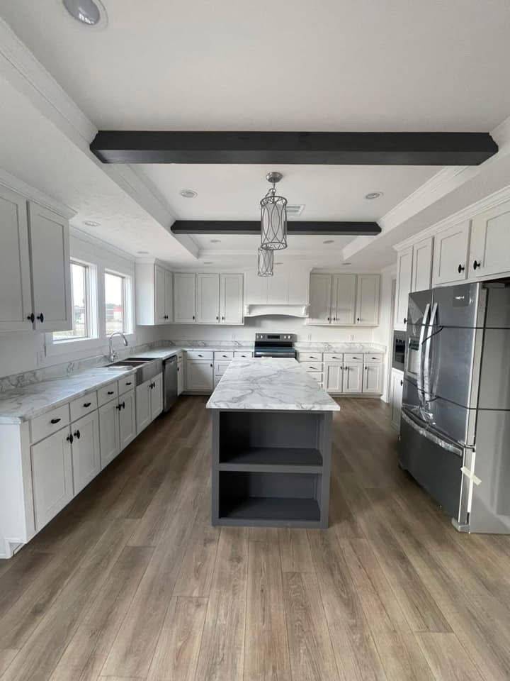 Spacious modern kitchen with white cabinets, marble countertops, central island, stainless steel appliances, wood floor, and dark ceiling beams. Inviting ambiance.