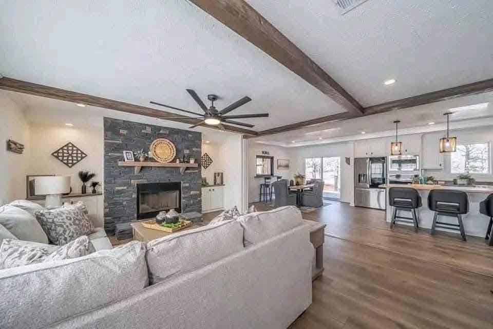 Spacious living room with neutral tones, featuring a gray stone fireplace, comfy sofas, wood ceiling beams, and open kitchen with bar stools. Cozy and modern.