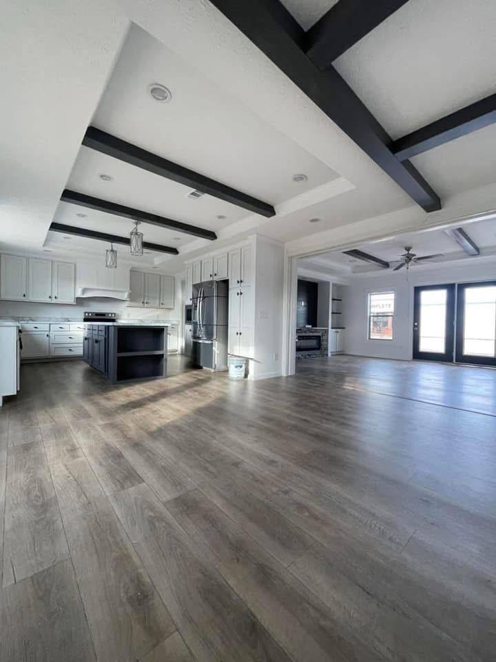 Modern kitchen and dining area with light wood floors, white cabinets, and a dark island. Black beams on the ceiling add contrast. Bright and spacious.