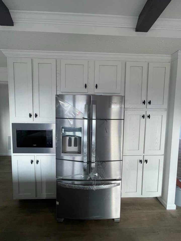 A modern kitchen with white cabinets creates a sleek backdrop for a large stainless steel refrigerator, complete with water dispenser. A microwave is built into the cabinets on the left. The room has a clean and organized feel.