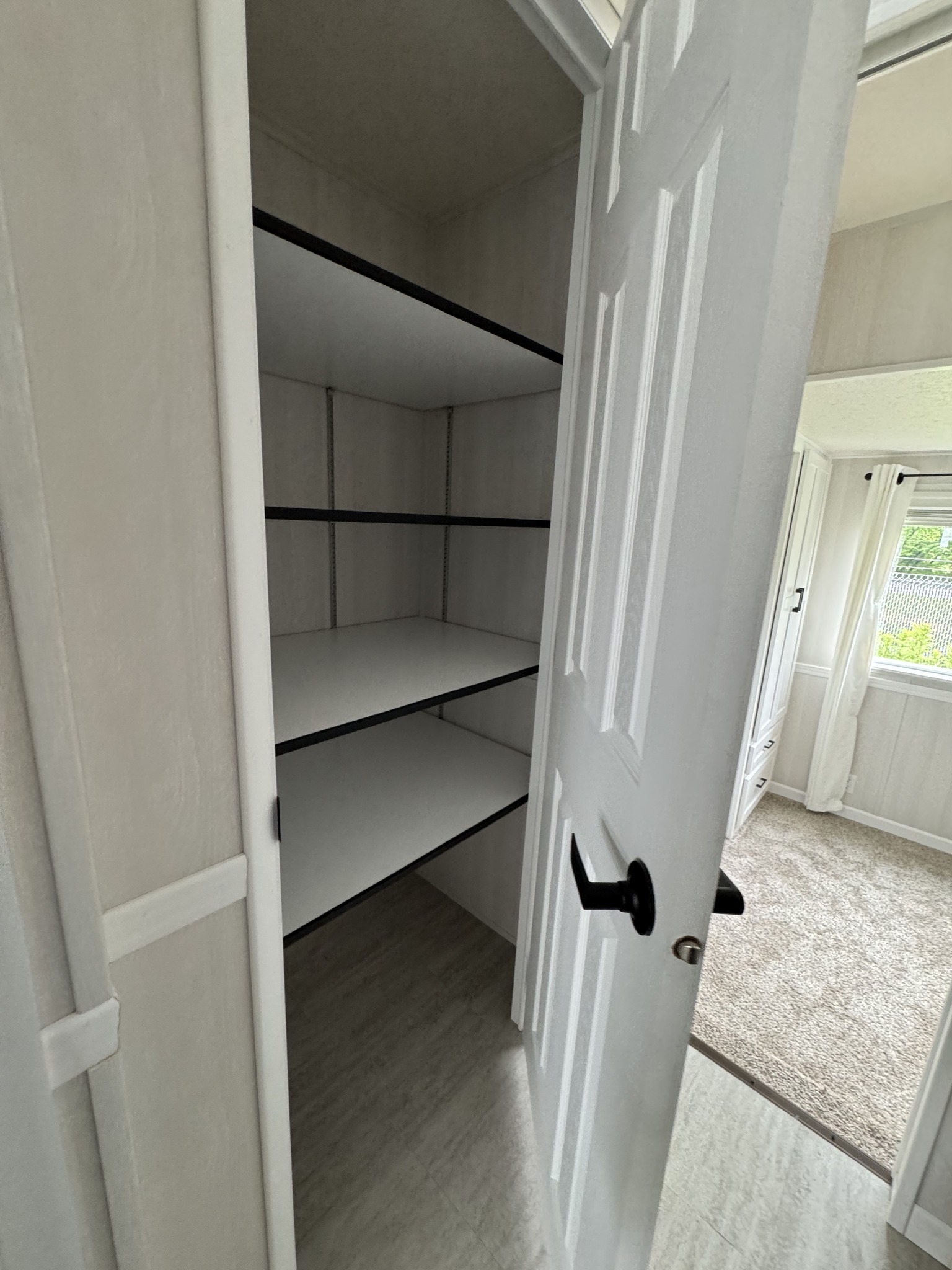 An open white door reveals an empty closet with four white shelves. The adjacent room has light-colored walls, carpet, and a bright window view.
