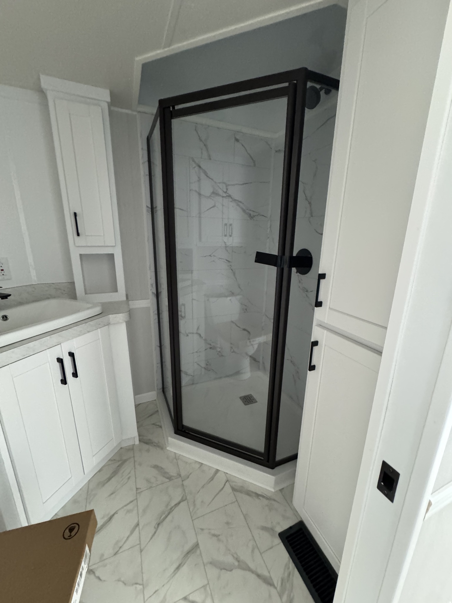 Compact bathroom featuring a modern glass shower with a black frame, white cabinets, and marble-patterned tiles on the floor and shower walls.