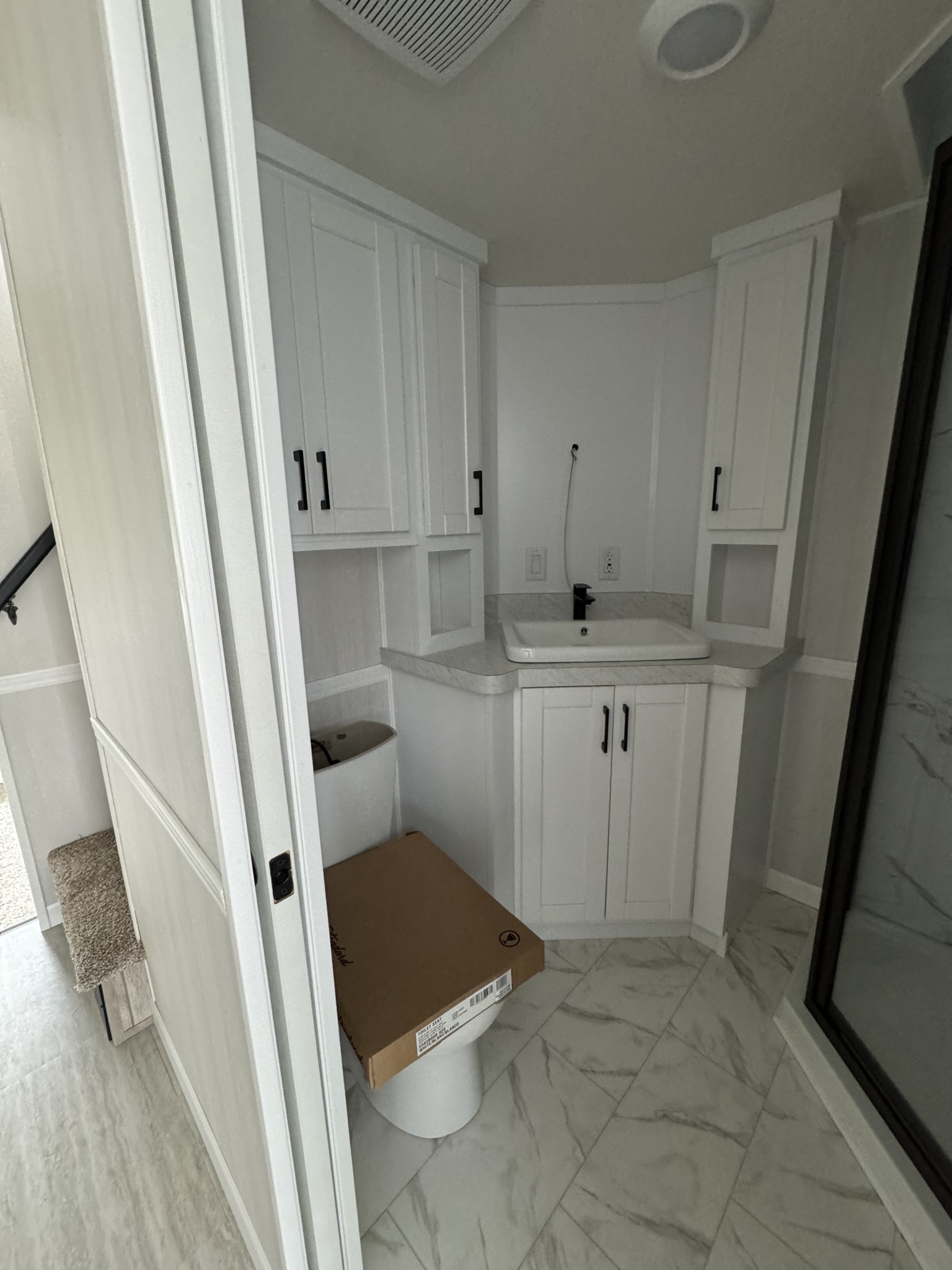 Compact, modern bathroom with white cabinets, a black faucet, and a sink. A cardboard box is on the toilet, and gray tile lines the floor.