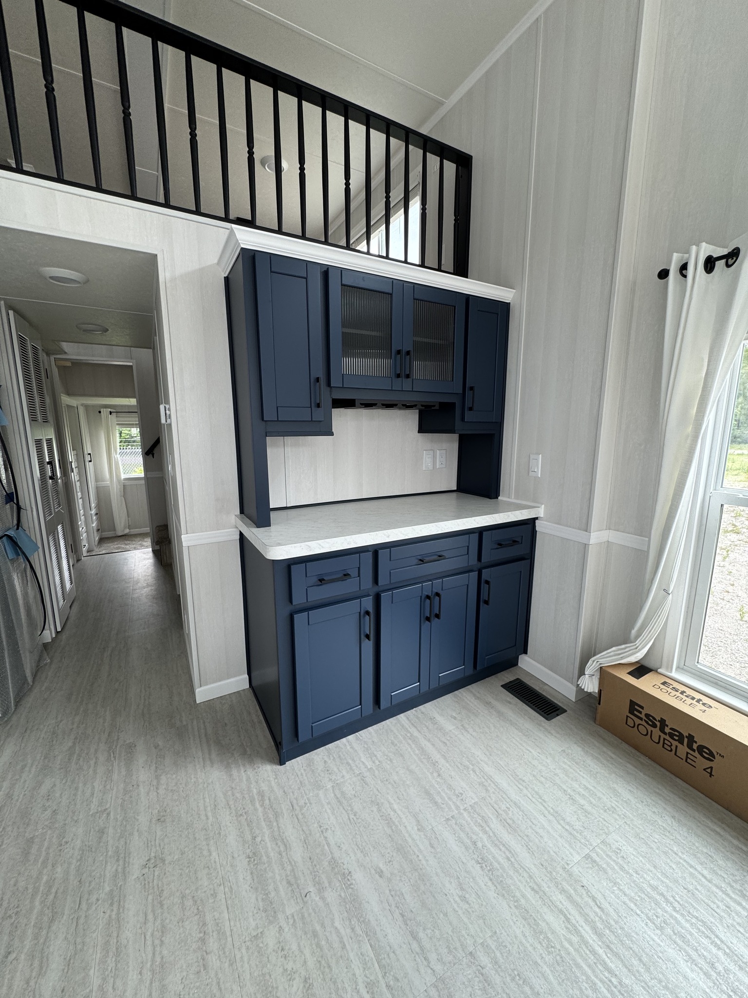Modern kitchen area with navy blue cabinets and white countertops under a loft with black railing. Bright, open atmosphere with light gray flooring.