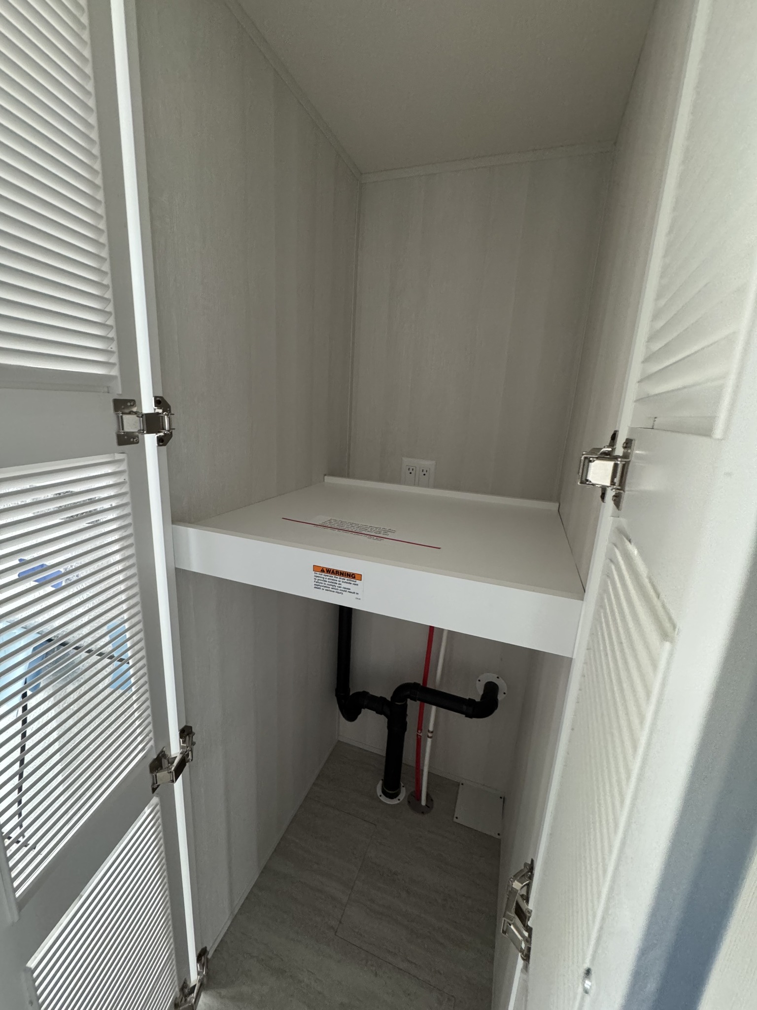 Small utility closet with white louvered doors open, inside a white shelf with electrical outlets and visible water pipes underneath, on a gray tiled floor.