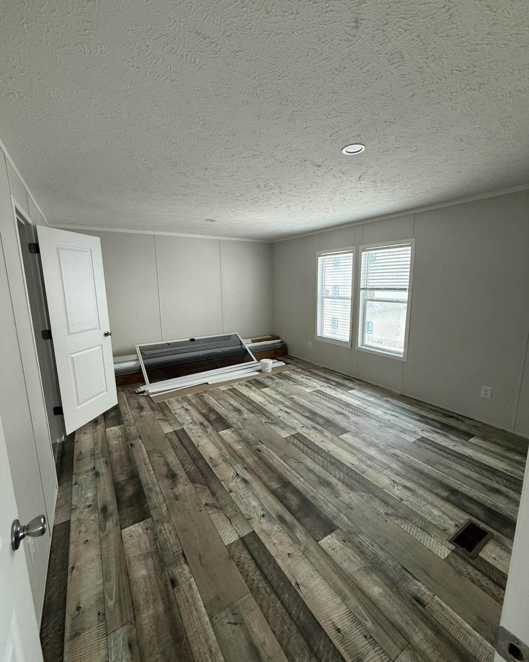Empty room with rustic wood flooring, white walls, and two windows letting in natural light. Disassembled furniture rests against one wall.
