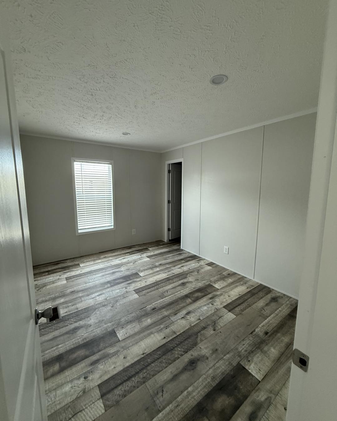 Empty room with rustic wood flooring, white walls, and a single window with blinds on the left. Dim lighting creates a calm, minimalistic atmosphere.