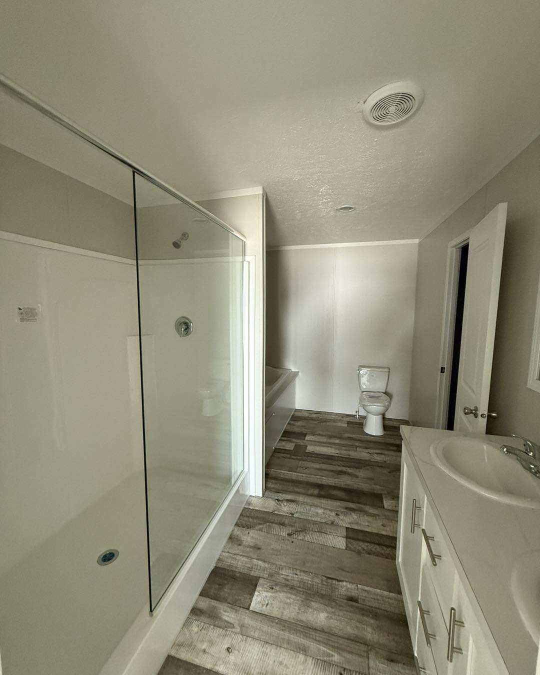 Modern bathroom with beige walls, wood-style flooring, and a glass-enclosed shower. Features double sink vanity, bathtub, and toilet, creating a clean, airy feel.