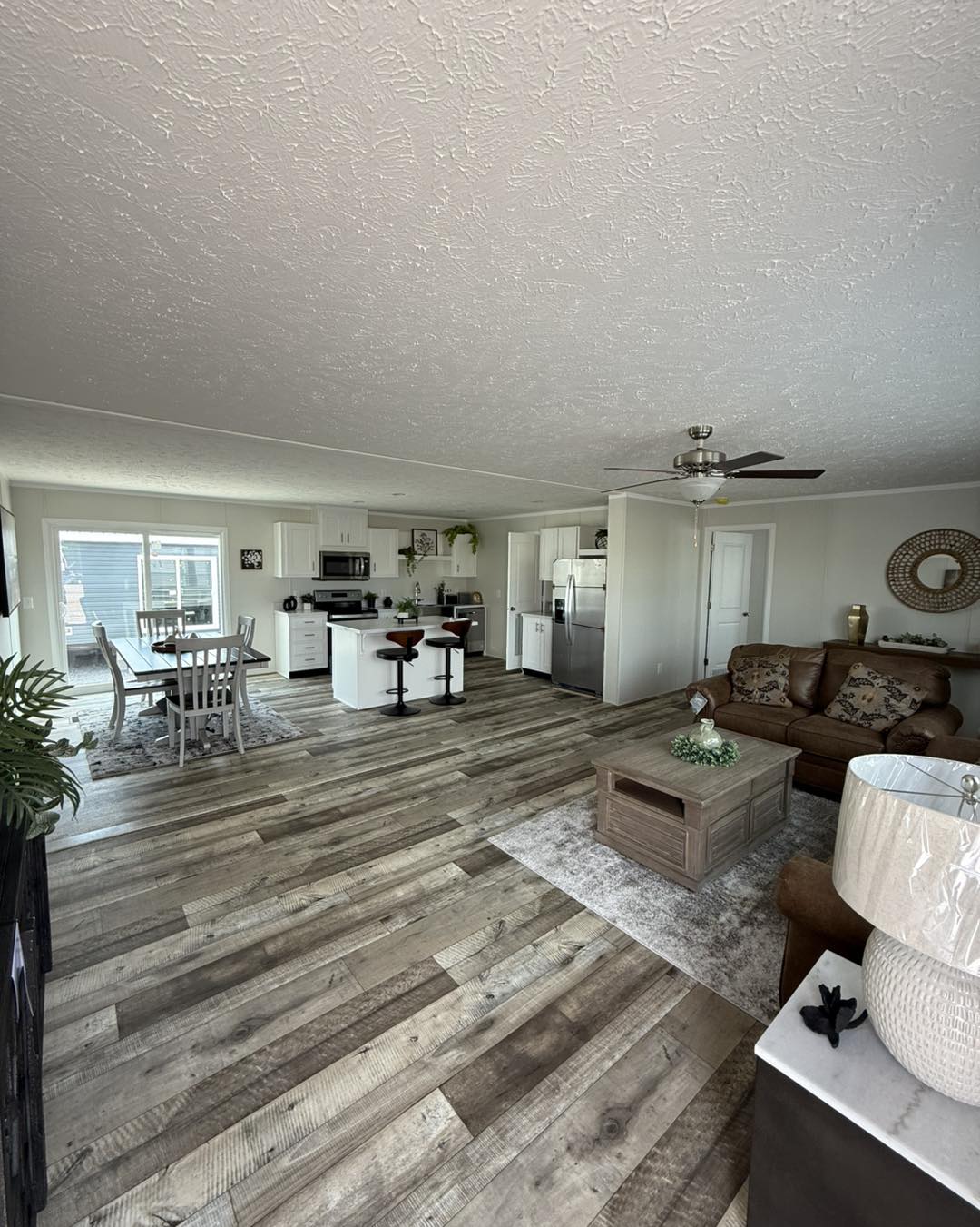 Spacious open-plan living room with wood floors, featuring a modern kitchen, island with bar stools, dining area by a window, and cozy seating with a brown sofa.