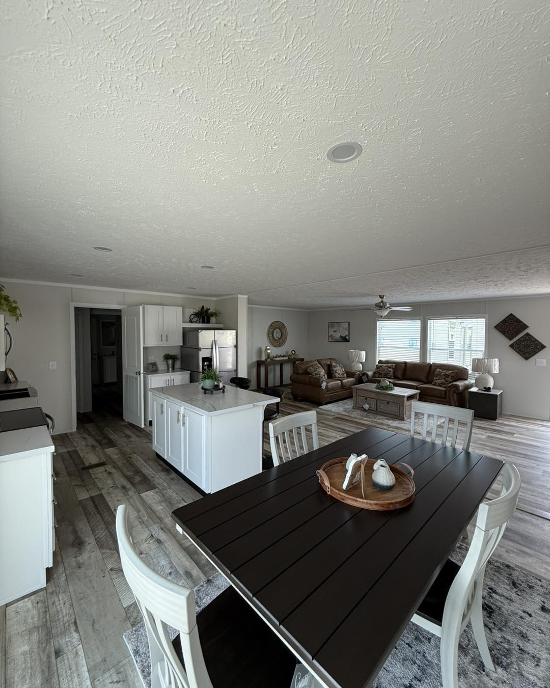 Open-plan living area with a modern kitchen and cozy lounge. Wood floors, a dark dining table, and a brown sofa set create a warm, inviting atmosphere.