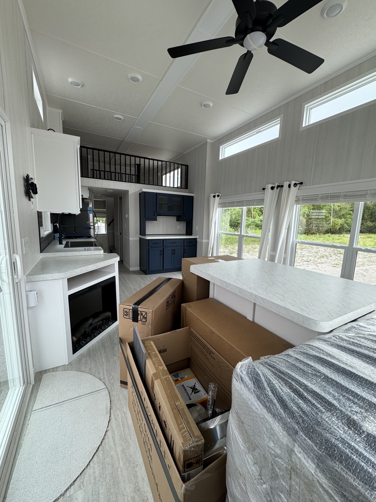 Interior of a modern, unfurnished room with white walls and ceiling, featuring a ceiling fan, large windows, and blue cabinets. Cardboard boxes are on the floor, conveying a new, moving-in vibe.