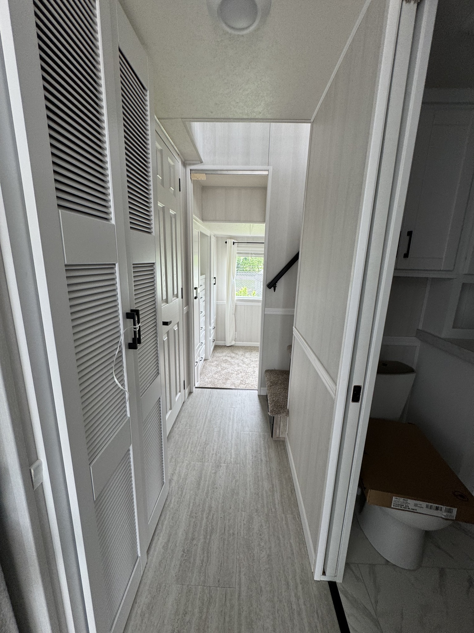 Narrow white hallway with light wood flooring. Open closet and visible toilet create a clean, compact atmosphere leading to a bright window.