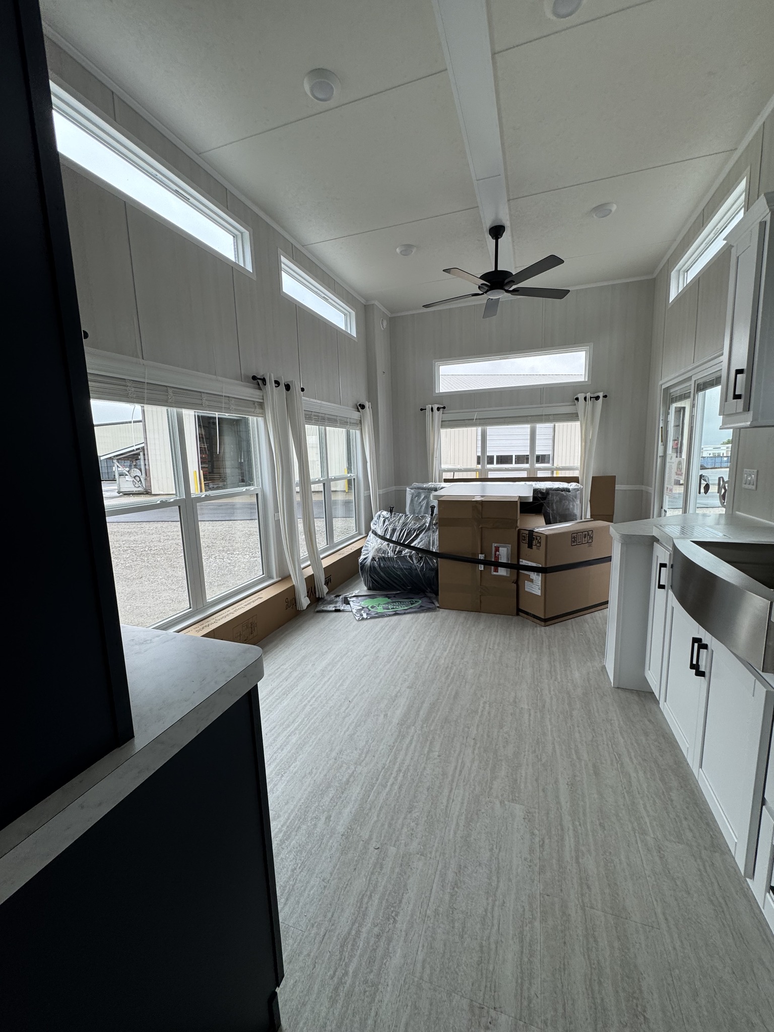 Minimalist modern room with high ceilings and large windows. Contains boxed furniture and a ceiling fan. Bright and airy atmosphere.