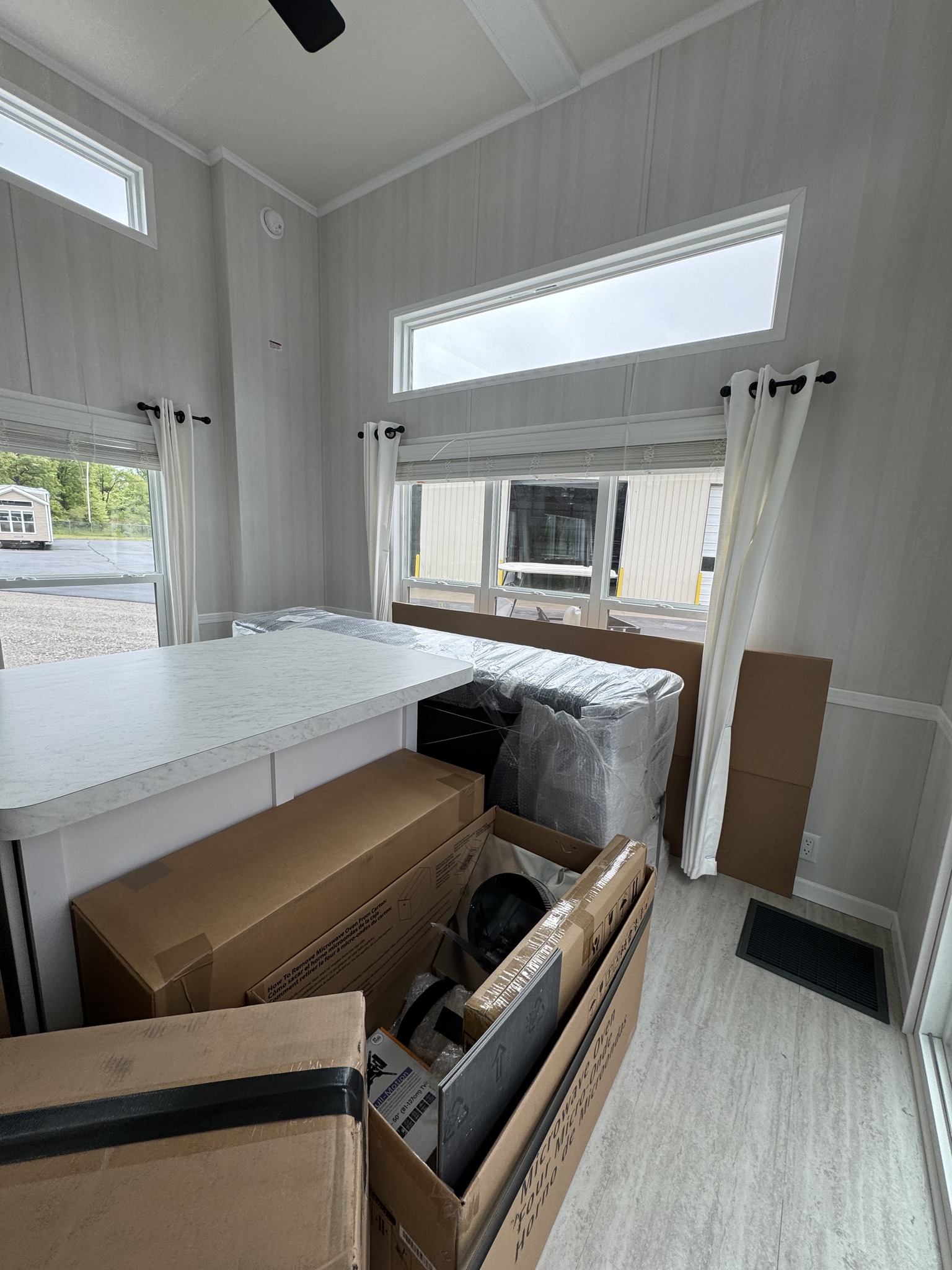 Interior of a small, brightly lit room with light wood paneling and multiple windows. The space is filled with unpacked boxes and plastic-wrapped furniture.
