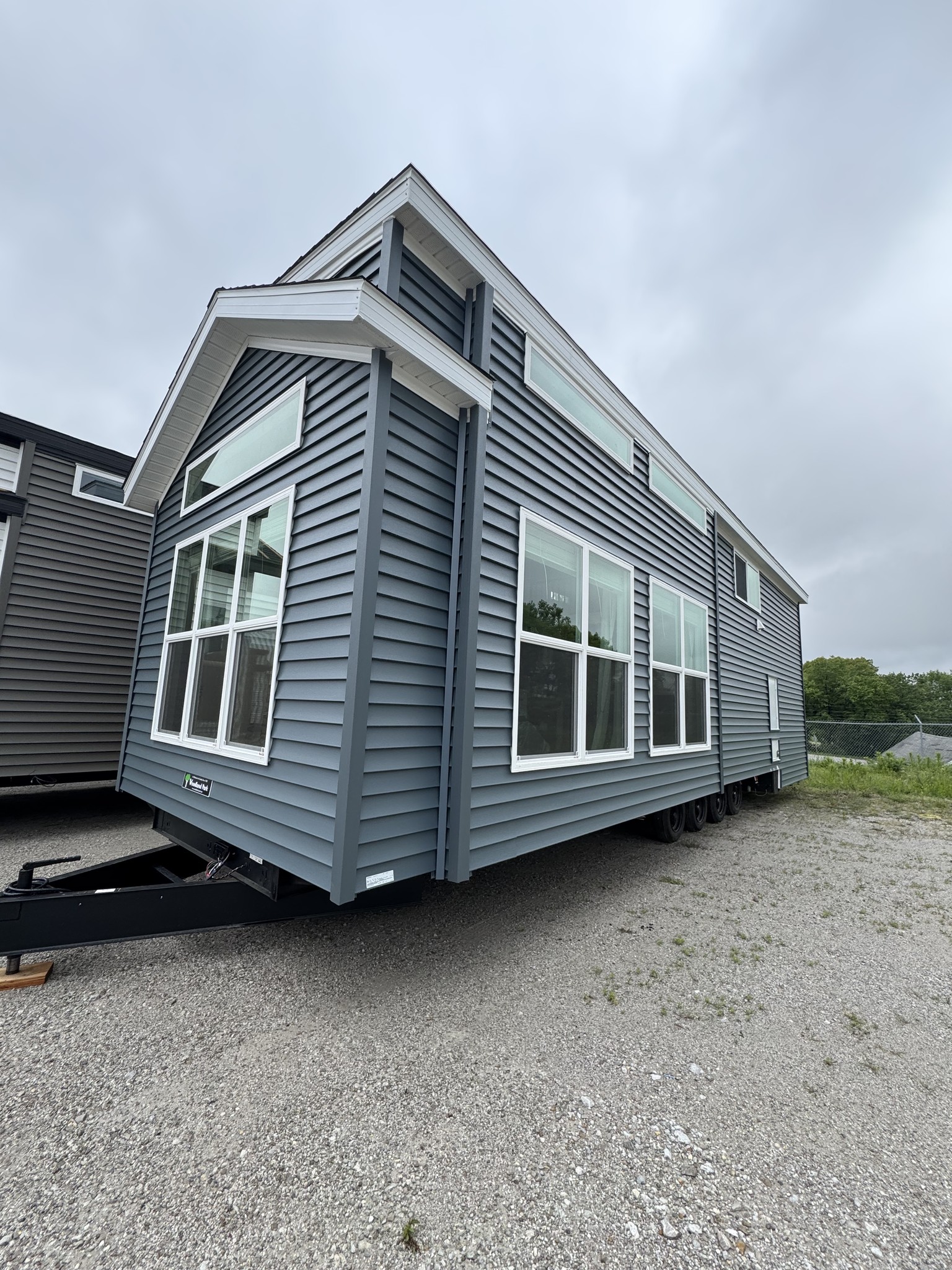 Modern tiny house on wheels with blue siding and large windows, parked on a gravel lot under an overcast sky, creating a cozy and minimalist vibe.