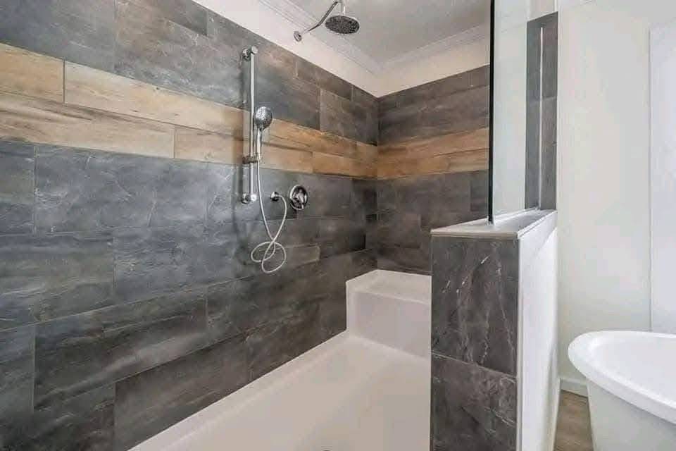 A modern bathroom features a sleek bathtub with a gray and wood-accented tile wall. There is a rain shower head and handheld shower, creating a spa-like feel.
