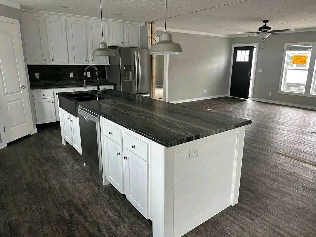 Modern kitchen with dark wood floor, white cabinets, and a black countertop island. Features stainless steel appliances and pendant lights. Bright, open space.