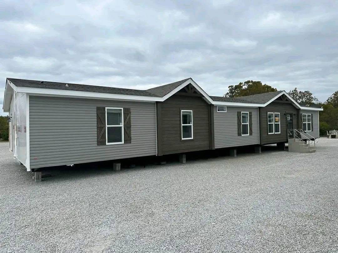 A large, gray manufactured home with a dark brown accent section and several windows sits on a gravel lot under a cloudy sky, conveying a tranquil atmosphere.
