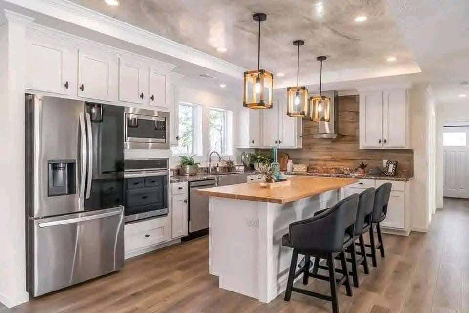 Modern kitchen with wooden floors, white cabinets, and a stainless steel fridge. An island features black chairs and pendant lights, creating a cozy ambiance.