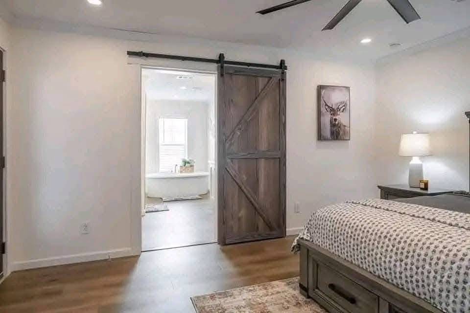 Modern bedroom with barn door leading to a bright bathroom. Features a cozy bed, a lamp on a nightstand, and a stag painting, evoking tranquility.