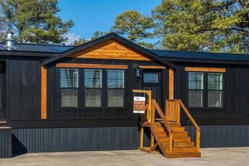 A modern black and wood-accented tiny house with a gabled roof sits on a concrete pad. It has a small wooden staircase and is surrounded by tall pine trees.