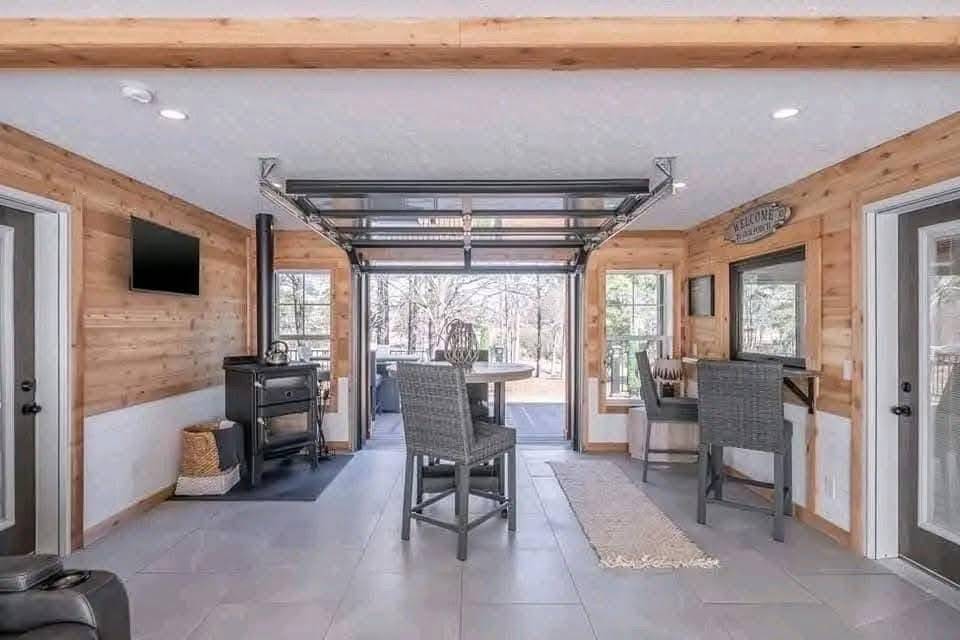 A cozy sunroom with wooden walls, a wood stove, high chairs around a round table, a TV, and a glass garage door opening to a bright view outside.