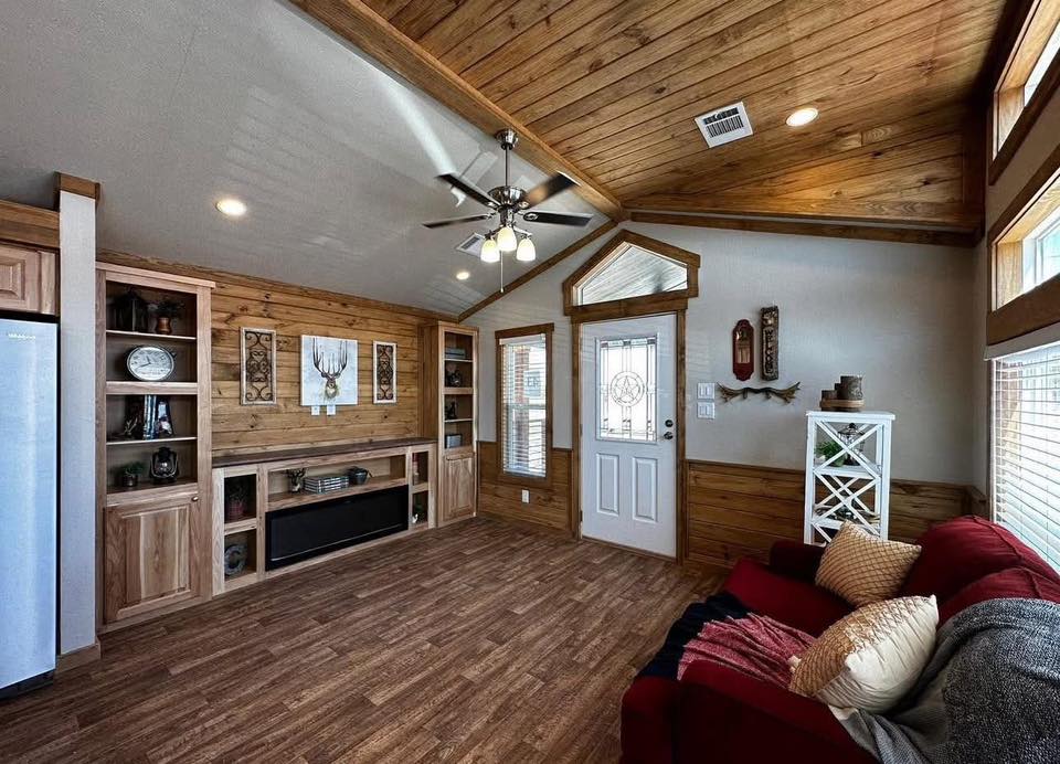 Cozy living room with wood-paneled walls and ceiling. It features a ceiling fan, shelves, a decorative door, red couch, pillows, and natural light.