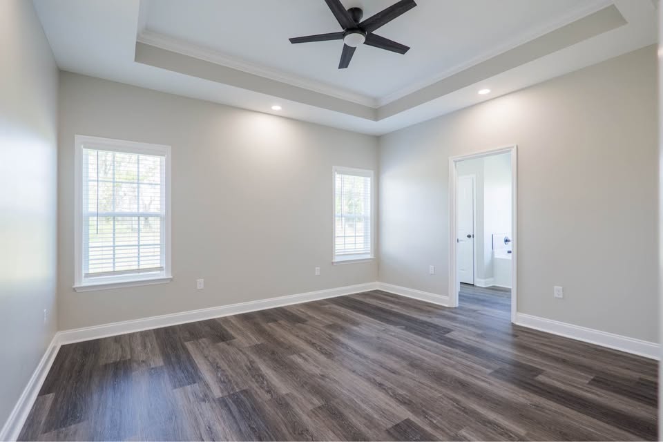 Bright, empty room with light gray walls, dark wood flooring, tray ceiling with a black fan, two windows, and an open door leading to a bathroom.