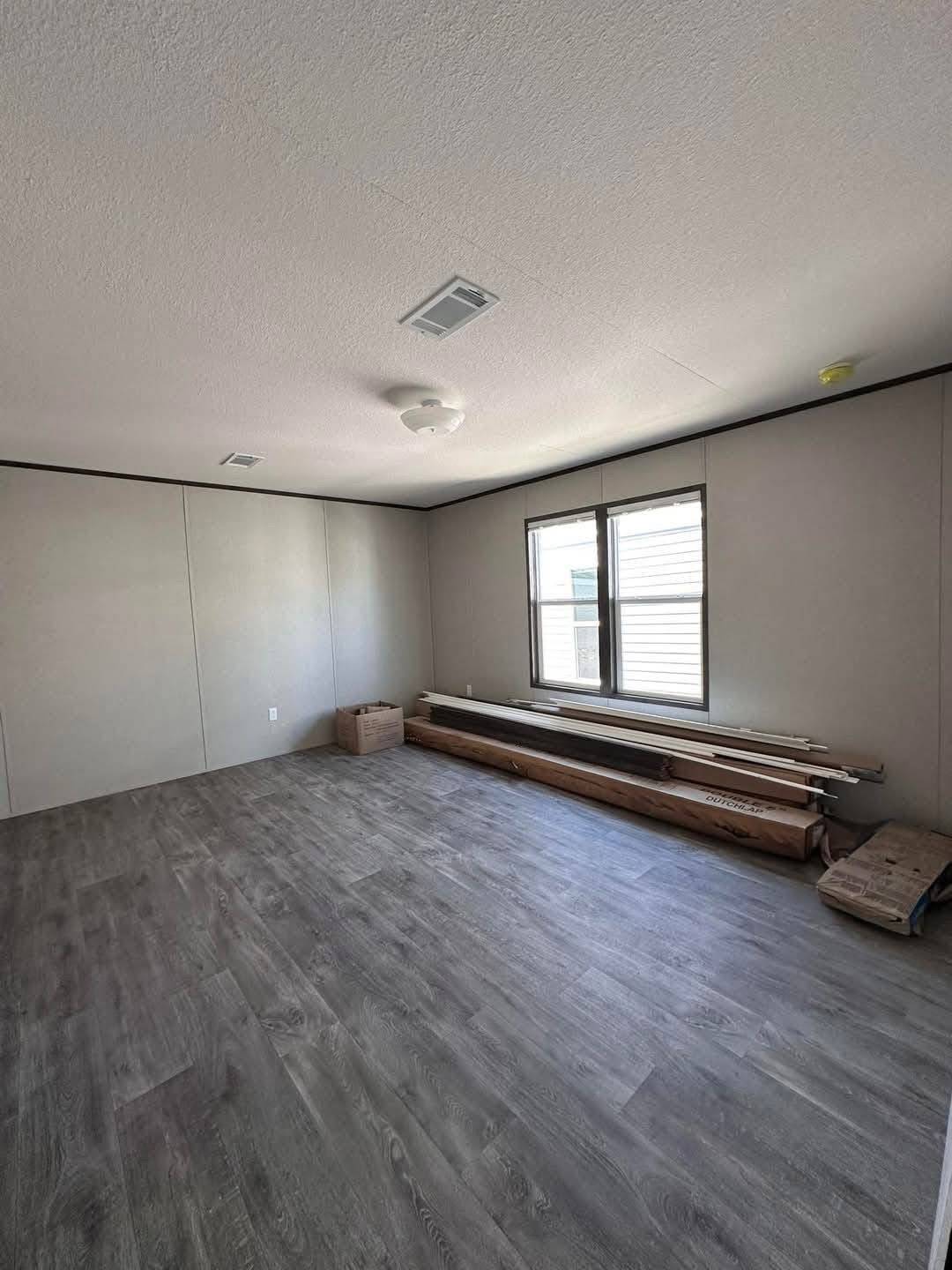 Empty room with light gray walls and dark wood flooring. A window on the right lets in natural light. Wooden planks and a cardboard box lie near the wall.