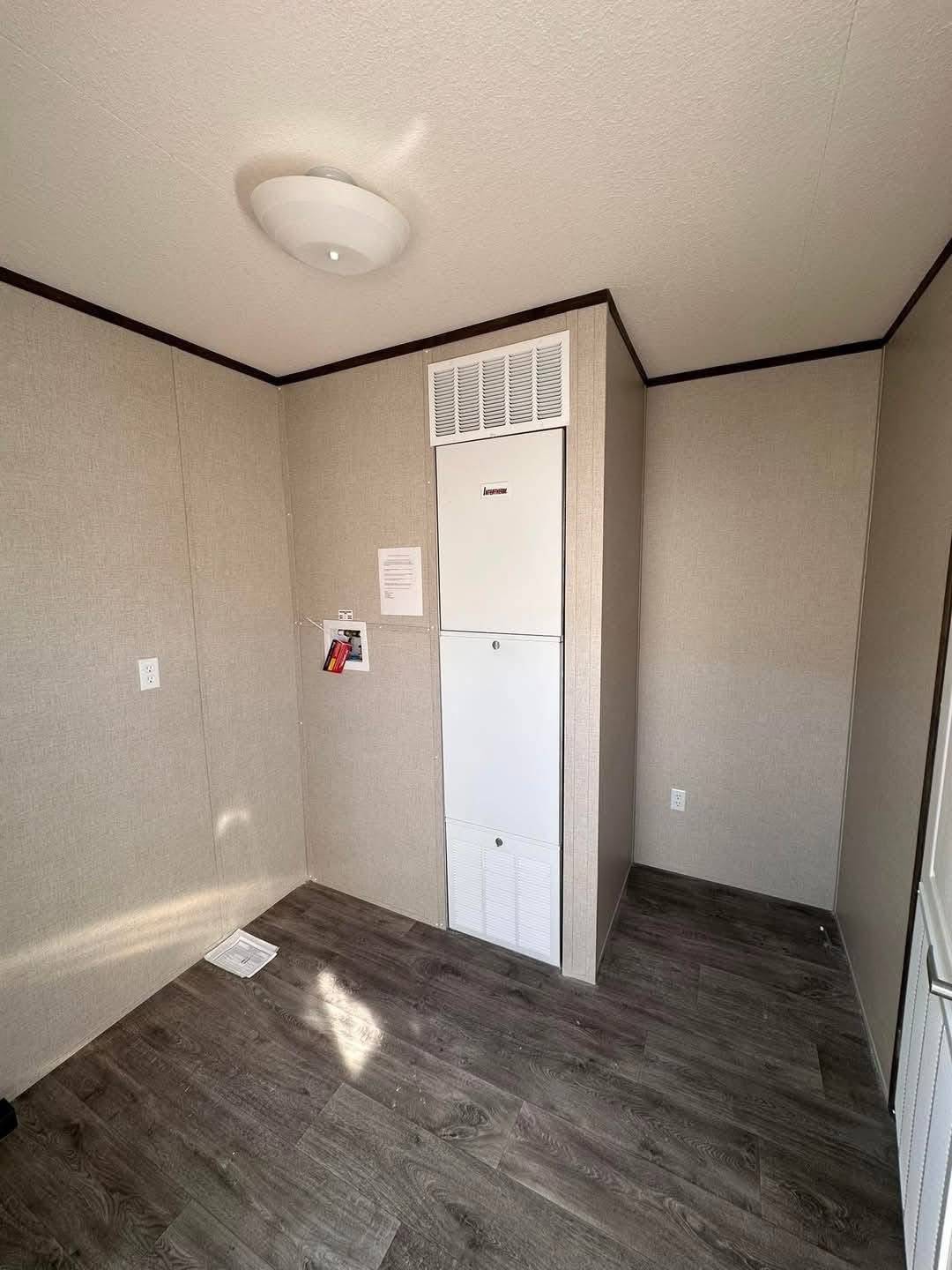 Simple room with light gray walls, dark wood floor, and ceiling light. A white HVAC unit with a vent is positioned against the corner wall. Minimalist and clean.