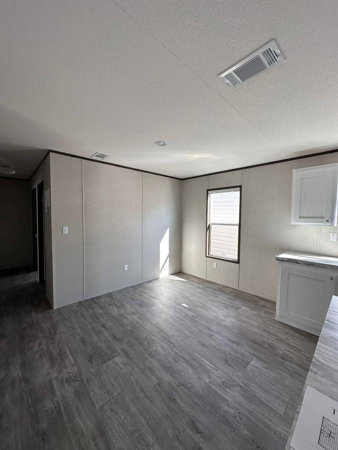Empty room with light gray walls, wood-look flooring, and a single window letting in sunlight. A small white cabinet is on the right, creating a minimalistic feel.