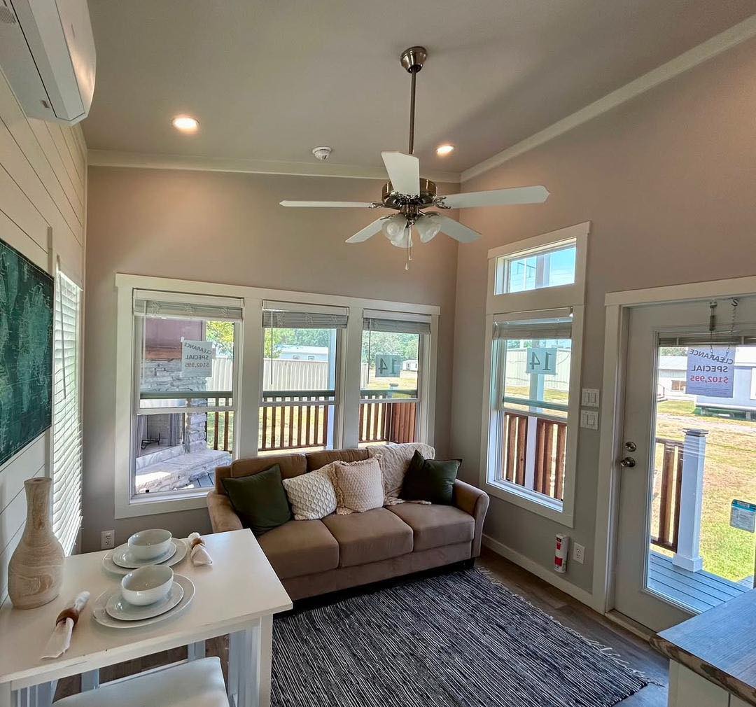 Cozy living room with a brown sofa, green and white pillows, under a ceiling fan. Bright windows and a doorway show an outdoor view. A small dining table with place settings is nearby, creating a warm, inviting atmosphere.