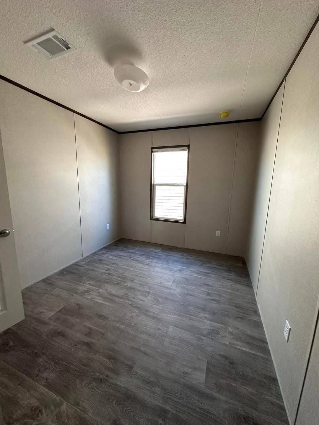 Empty room with beige walls and dark wood flooring, featuring a centered window with blinds. Ceiling light fixture provides a minimalistic feel.