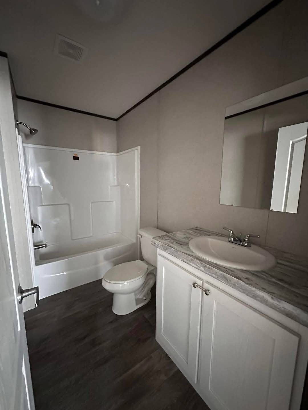 Modern bathroom with dark wood flooring, featuring a bathtub with shower, toilet, and a sink on a marble countertop beside a mirrored cabinet.