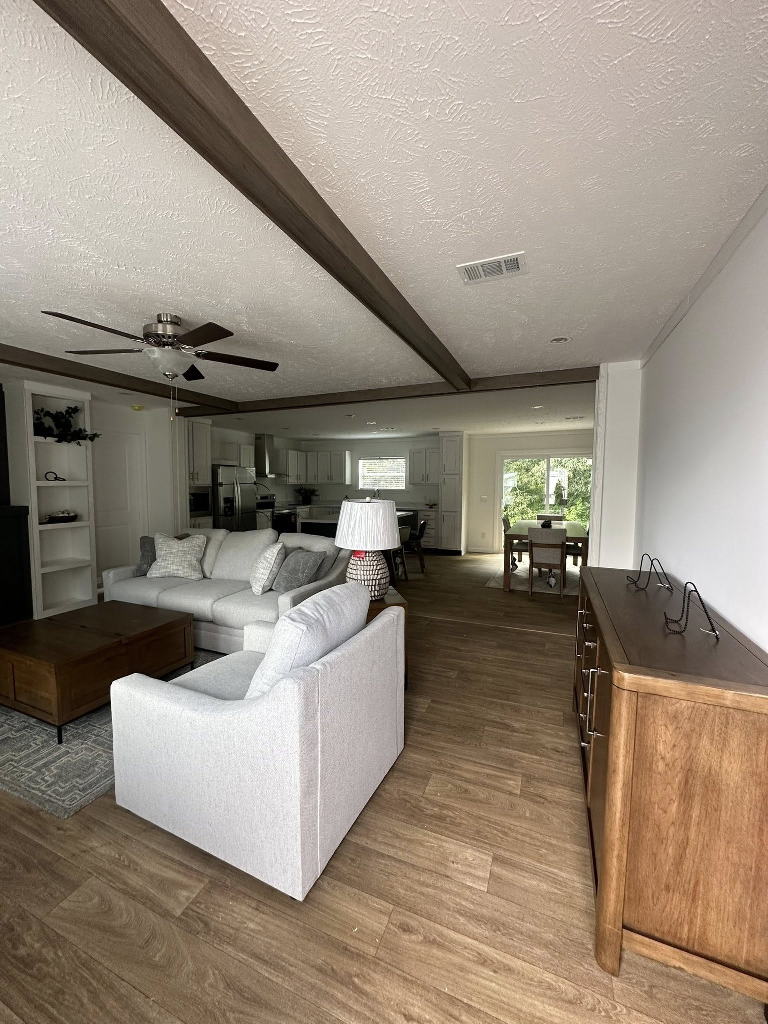 Spacious living room with neutral tones, featuring a gray sofa, wooden ceiling beams, and a modern fan. Open layout leads to kitchen and dining area.