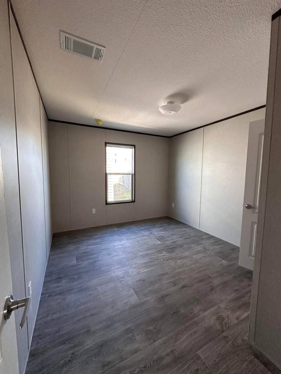 An empty room with light gray walls and a textured white ceiling, featuring a large window and dark wood laminate flooring, creating a simple, clean, and bright atmosphere.