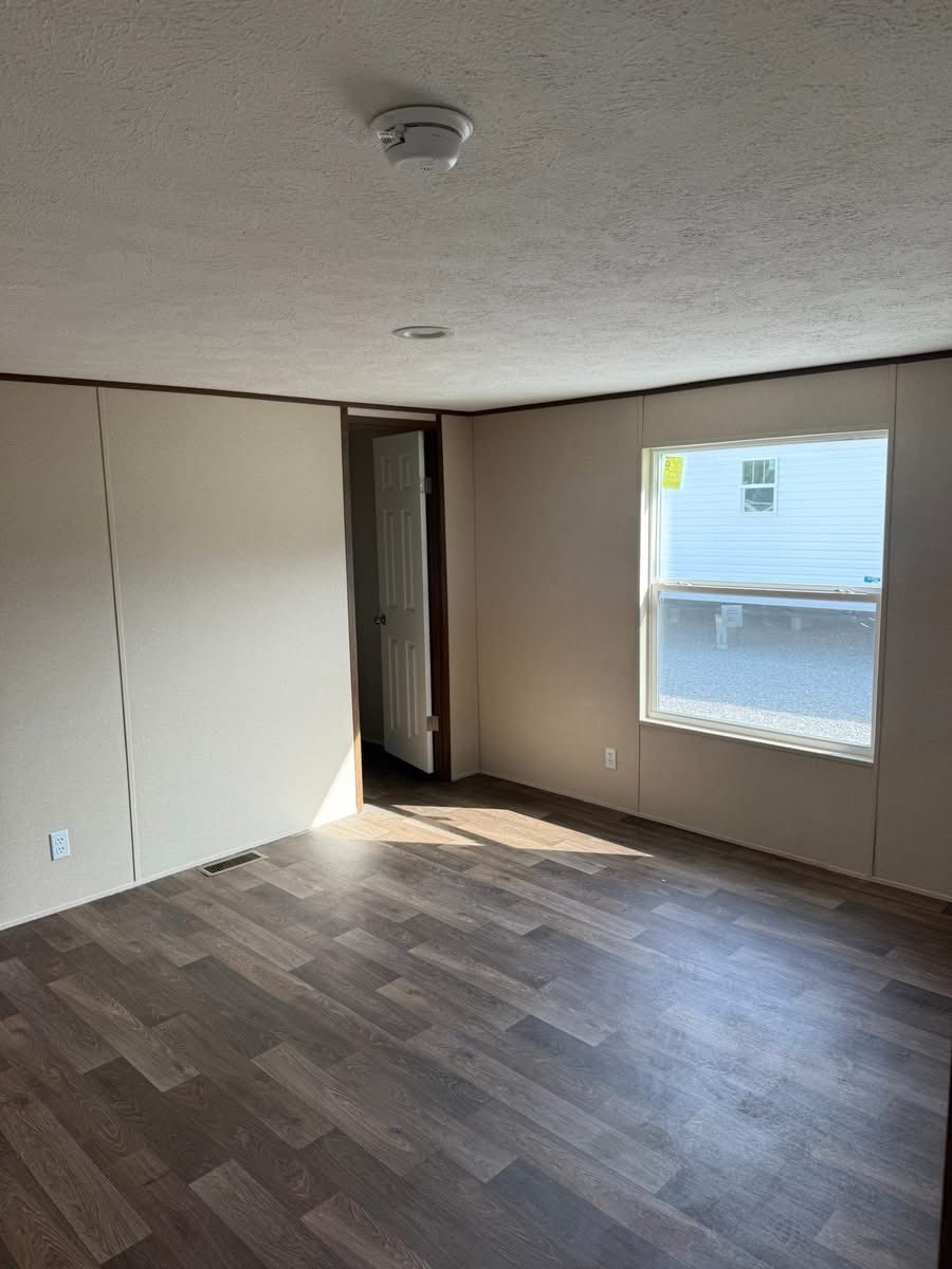 Empty room with tan walls and wood-patterned floor. A window on the right lets in light, and there's a partially open door in the corner. Quiet, simple ambiance.