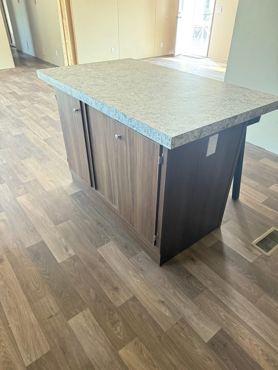 A wooden kitchen island with a light gray countertop stands on brown laminate flooring. The room appears bright, with neutral wall tones, creating a modern, minimalist feel.