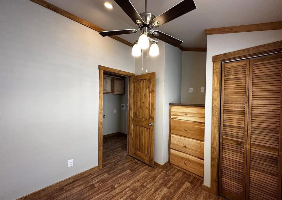 A small room with wooden flooring and trim features an open wooden door leading to a closet. A ceiling fan with lights hangs from above, and a wooden dresser is beside a louvered closet door.